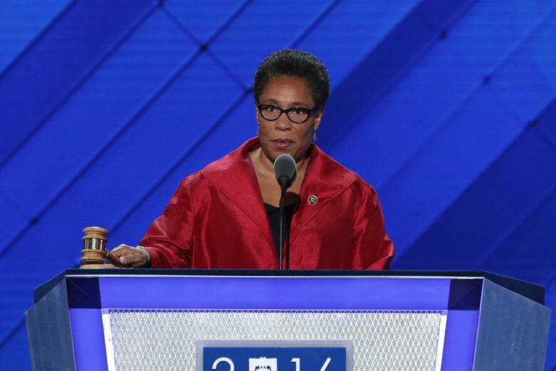 Marcia Fudge, dressed in red, holds a gavel at a podium with "2016"—the "0" replaced by a drawing of the Liberty Bell—on the front. The backdrop and the podium are both blue.